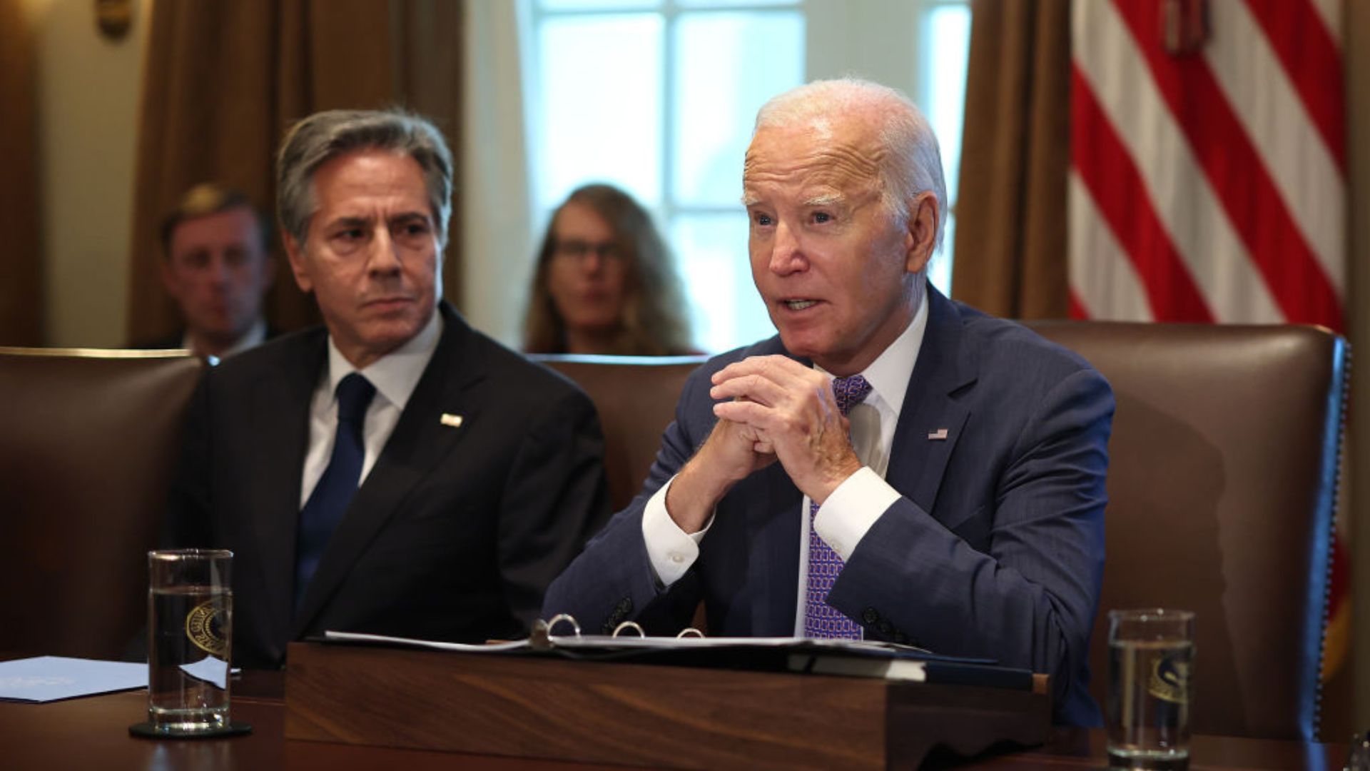 Joe Biden and Antony Blinken seated at a wooden table in a room with draped windows and an American flag in the background