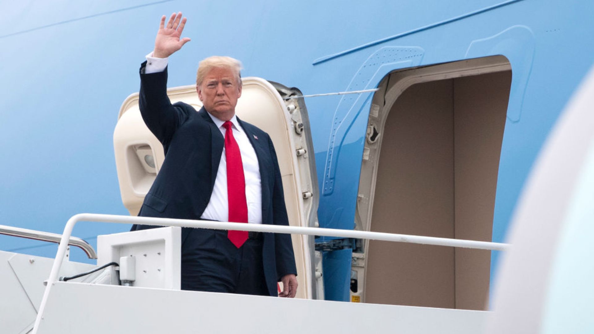 Donald Trump standing at the doorway of Air Force One, raising his right hand in a wave
