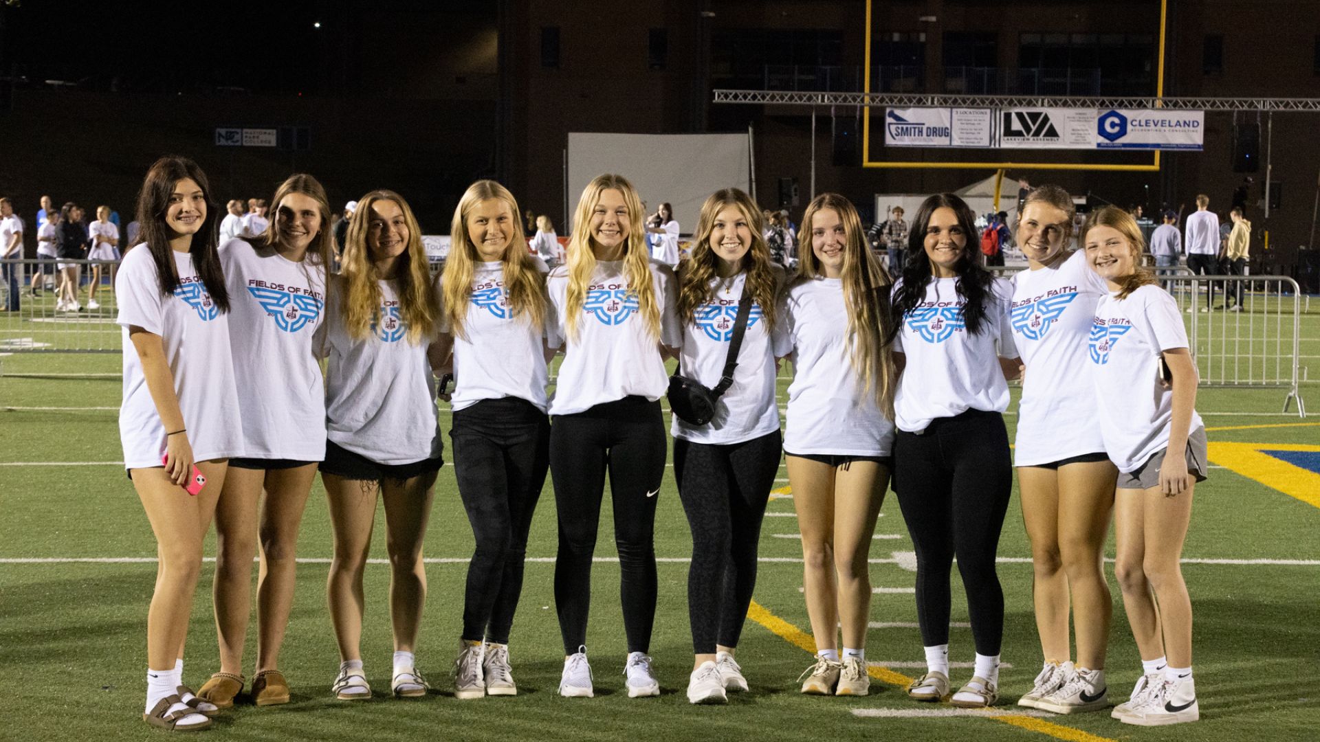 A group of young women standing together on a football field at night, all wearing white t-shirts with "FIELDS OF FAITH" and a blue graphic design
