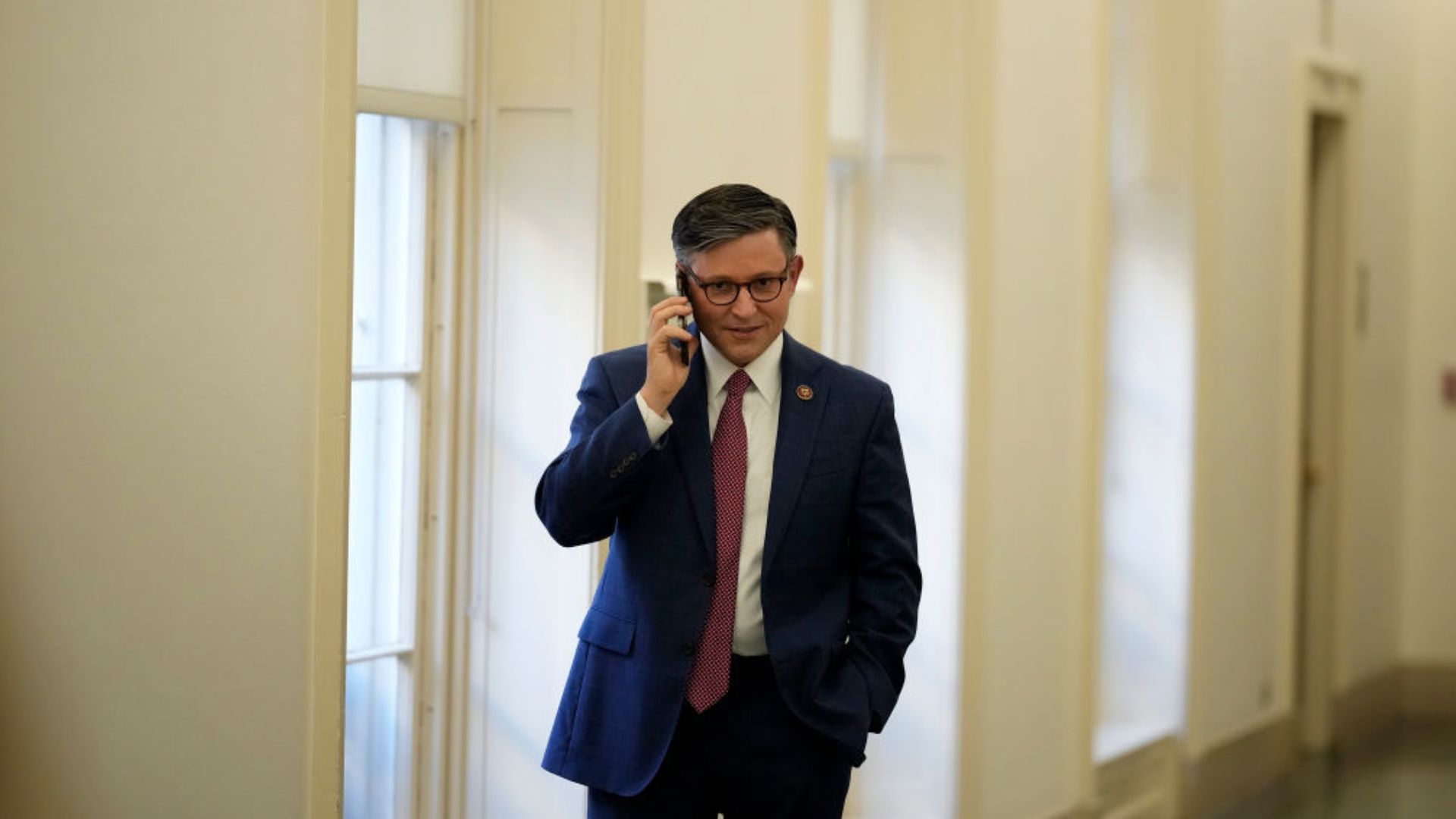 Rep. Mike Johnson, wearing a blue suit, white shirt, and red tie, is seen speaking on a mobile phone. He has glasses on and stands in a hallway with white walls and doors.
