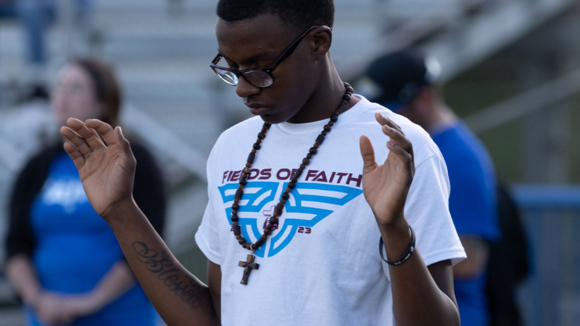 A young man wearing glasses, a white t-shirt with "Fields of Faith" printed on it, and a beaded necklace with a cross pendant, is captured in a moment of reflection or prayer