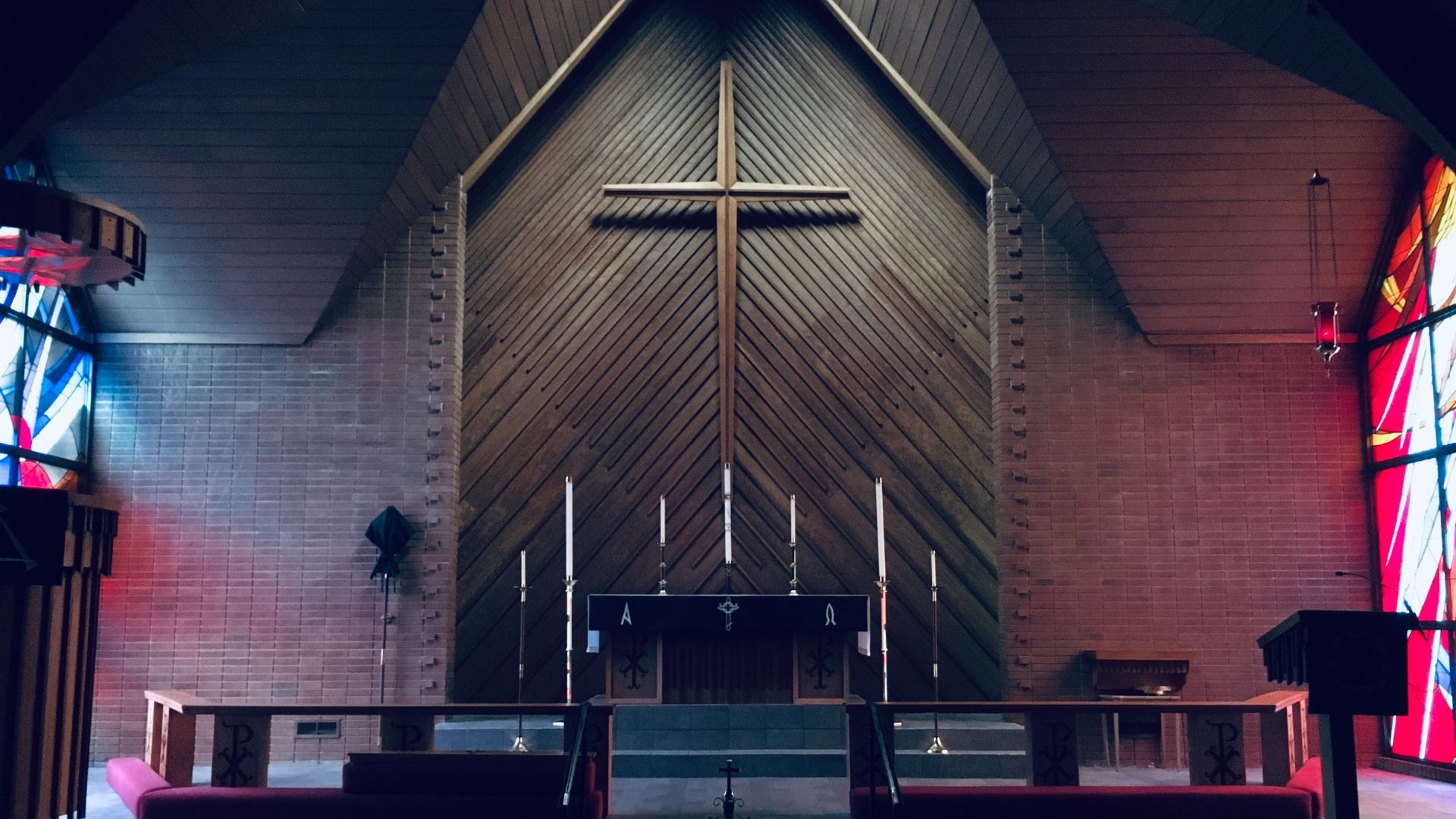 A symmetrical view of a church interior featuring a large wooden cross mounted on a slanted wooden wall. On either side, there are stained glass windows casting colored light onto the brick walls