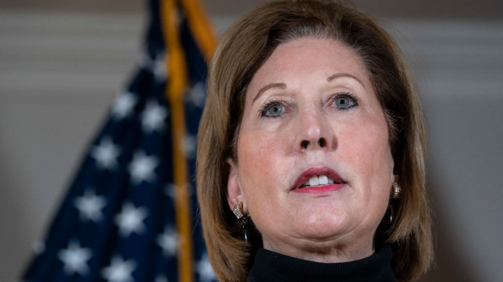 Sidney Powell, an attorney, appearing in a public setting. She is pictured up close, with an American flag partially visible in the background