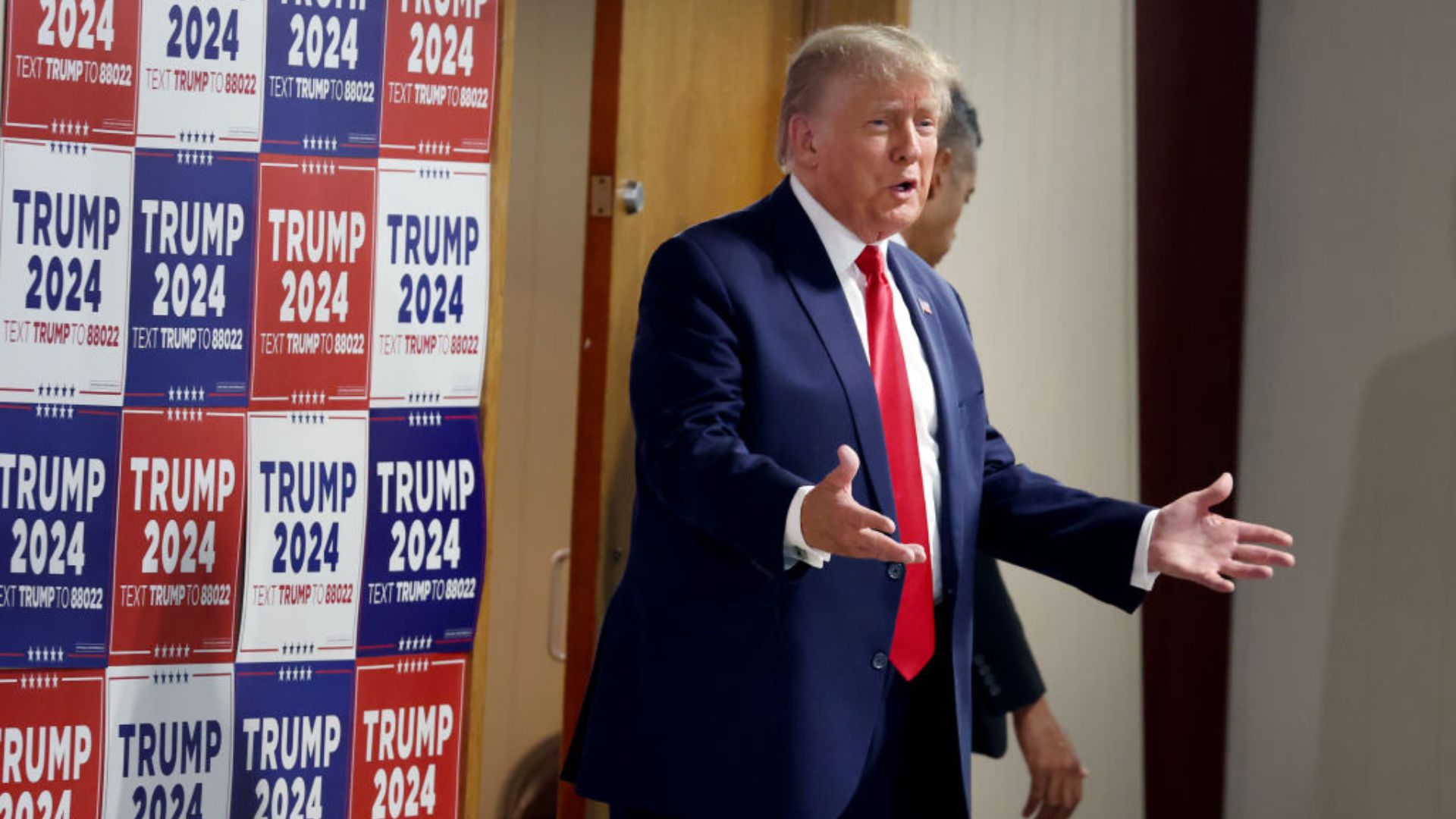 Donald Trump, dressed in a dark blue suit with a red tie, gestures with his right hand while speaking. Behind him are multiple vertical banners showcasing 'TRUMP 2024' in red and blue colors, with the text 'TEXT TRUMP TO 88022' below each