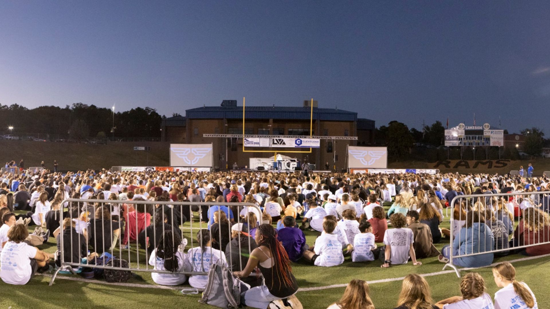 A vast number of people sitting and standing on a football field in the evening, facing a distant stage with various banners