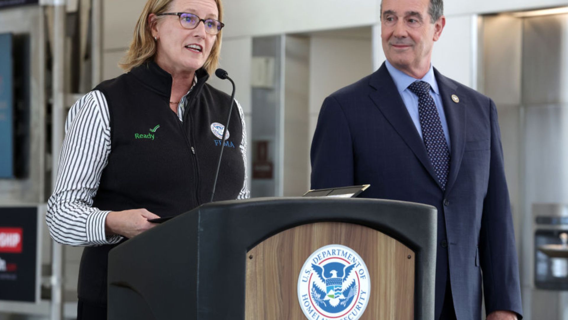 A woman wearing glasses, a striped shirt, and a black vest labeled 'Ready' and 'FEMA' speaks into a microphone at a podium with the U.S. Department of Homeland Security emblem