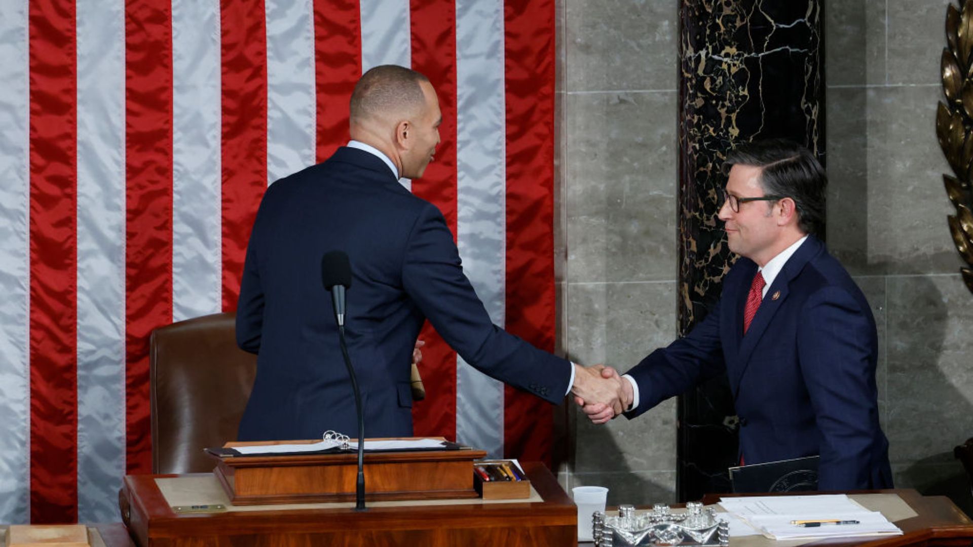 Rep. Mike Johnson, in a blue suit and wearing glasses, is seen seated to the right, shaking hands with a bald man in a darker suit standing to his left. They are in front of an American flag backdrop.