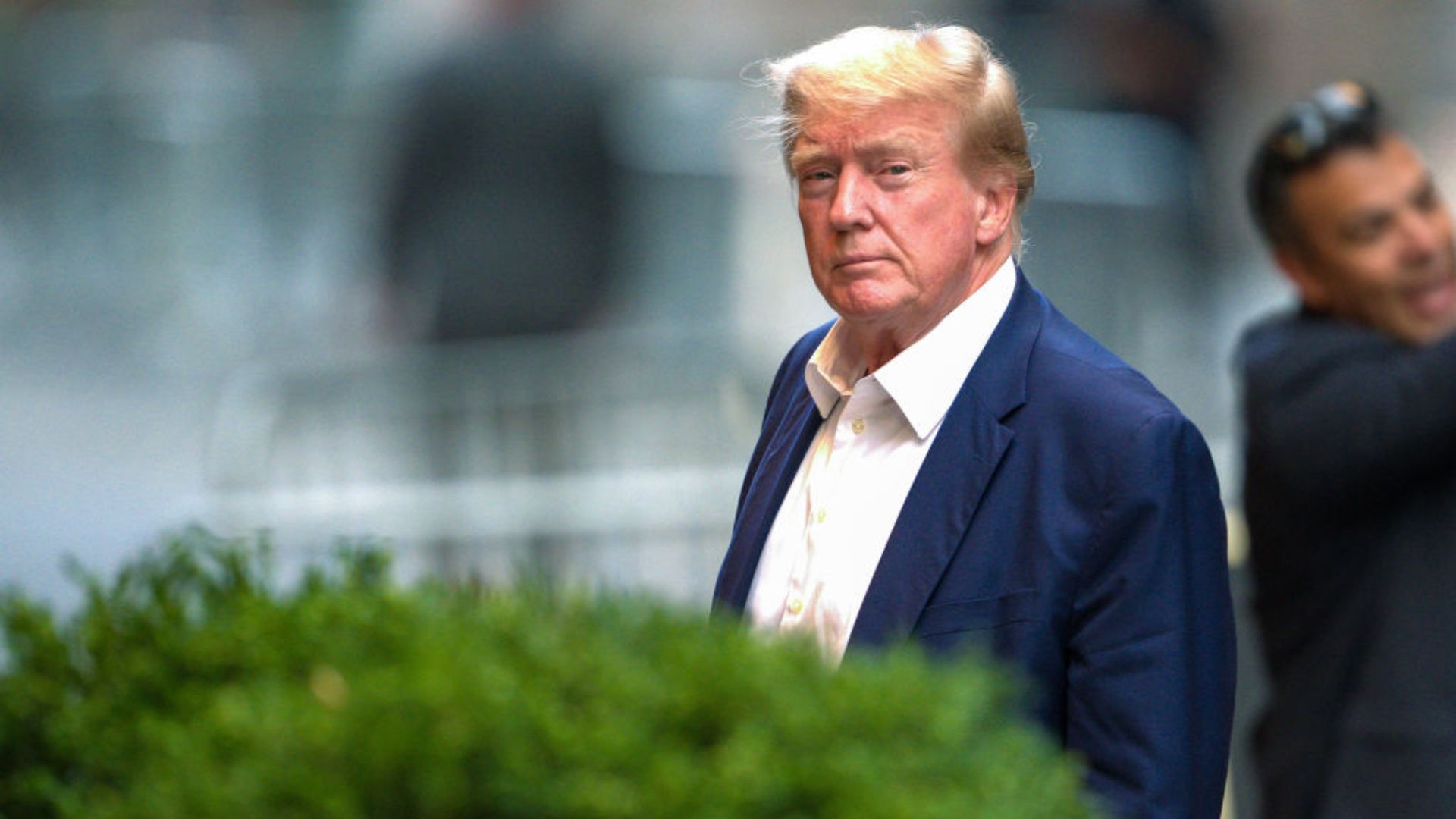 Donald Trump, wearing a navy suit and white shirt, standing outdoors with an urban backdrop. He has a serious expression, and there's a blurred individual with sunglasses in the background