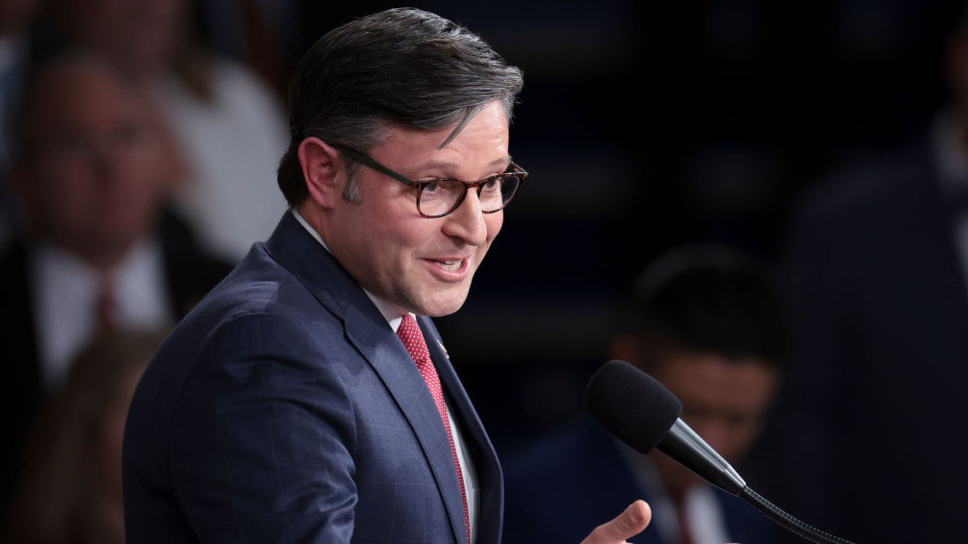 Rep. Mike Johnson in a blue suit and red tie, wearing glasses, speaking into a microphone with a focused expression, with blurred attendees in the background