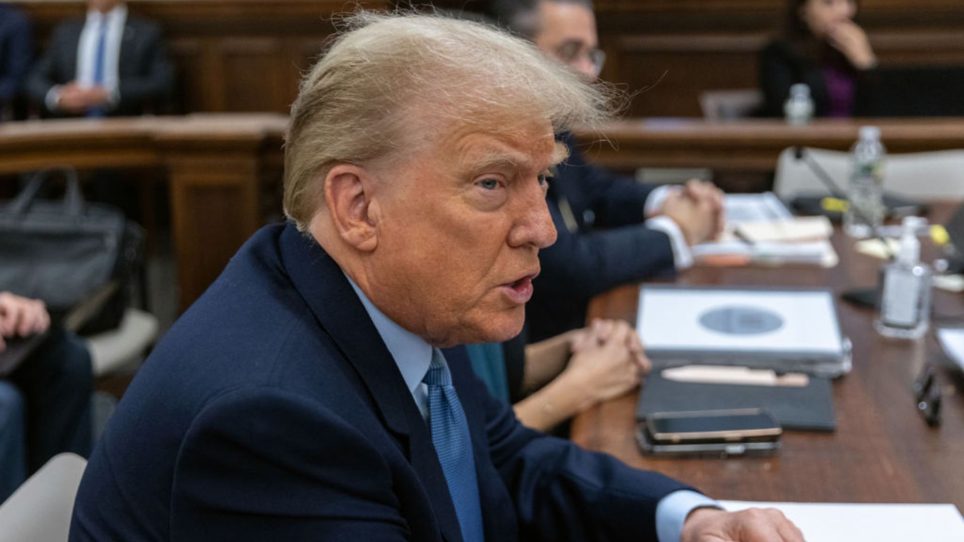 Former President Donald Trump participating in a meeting, captured in mid-speech, surrounded by various attendees and meeting materials