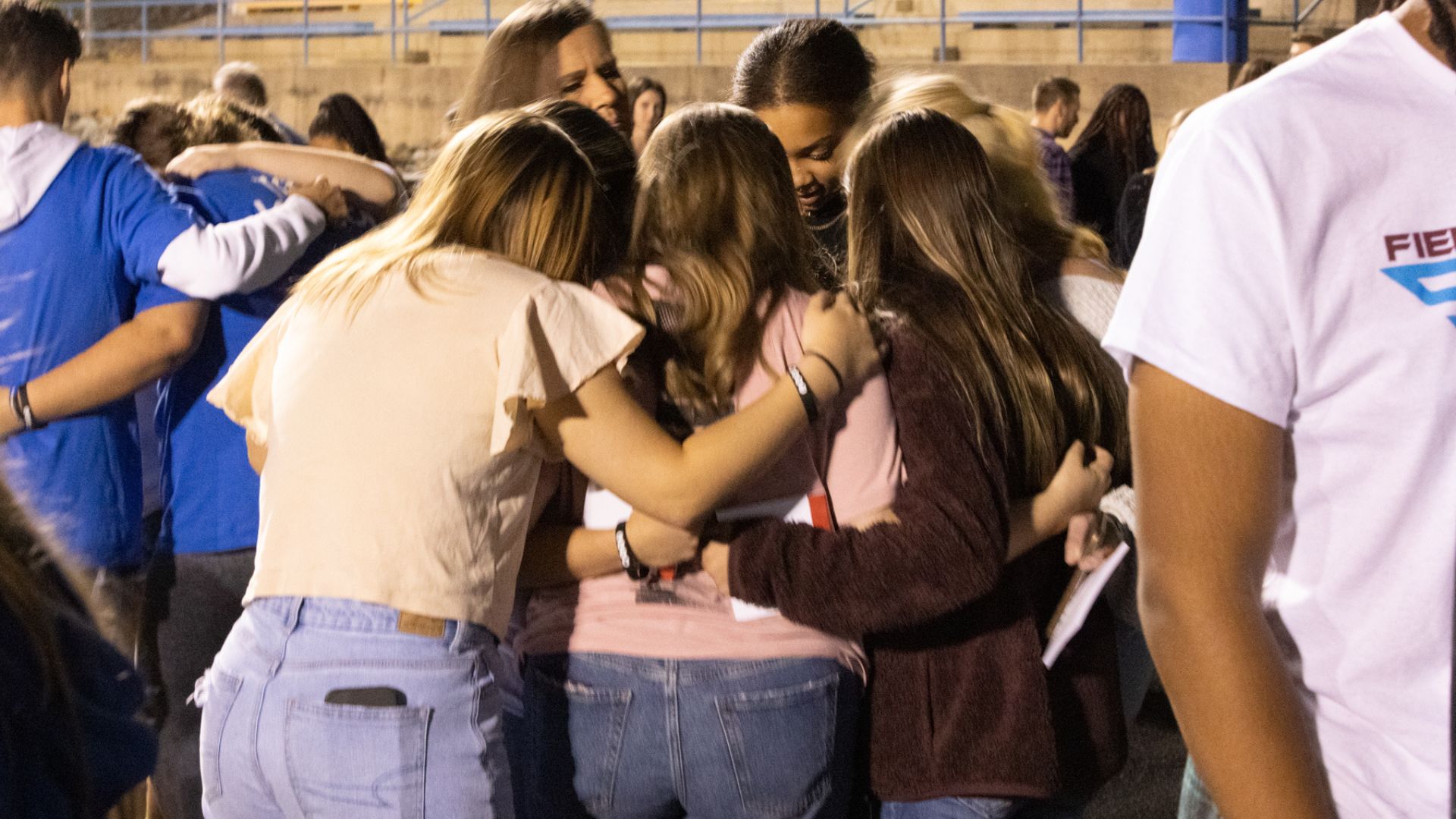Several individuals at an evening event, forming a close-knit group hug. The group mainly consists of young people wearing casual attire like jeans, t-shirts in various colors, and wristbands.