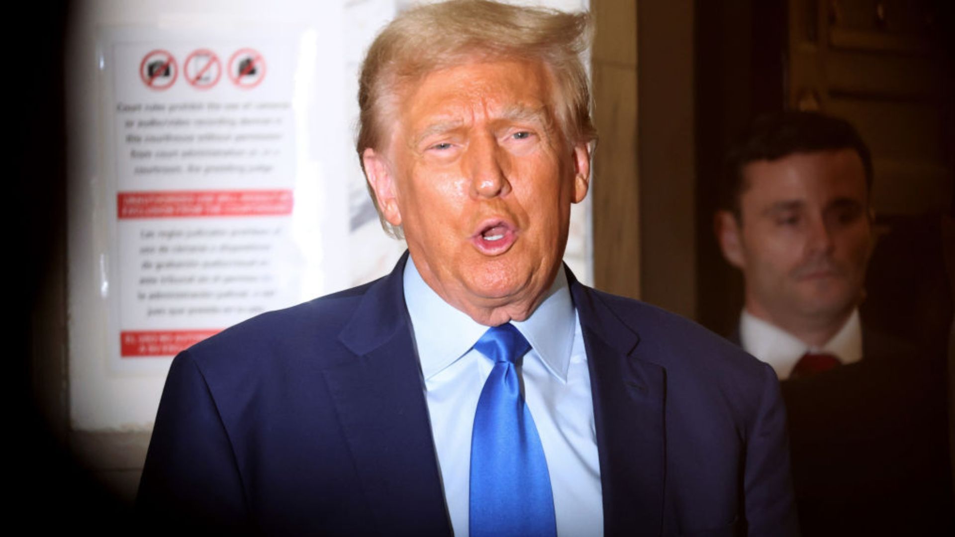 Former President Donald Trump, wearing a blue suit and tie, is captured mid-sentence in a photograph