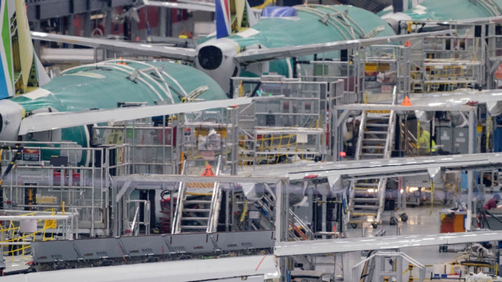 A close-up view of an aircraft assembly line. Multiple partially constructed airplanes with green protective coatings are surrounded by metal scaffolding