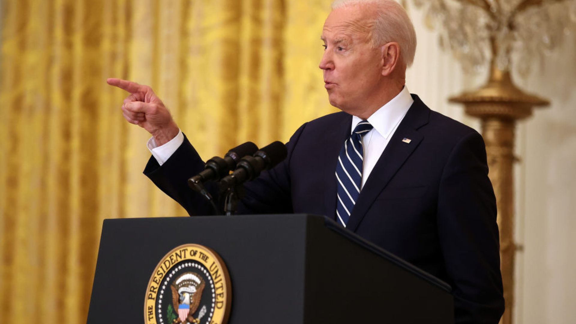 U.S. President gesturing with his right hand while speaking into microphones at a podium with the presidential seal, set against a backdrop of golden curtains and a decorative lamp