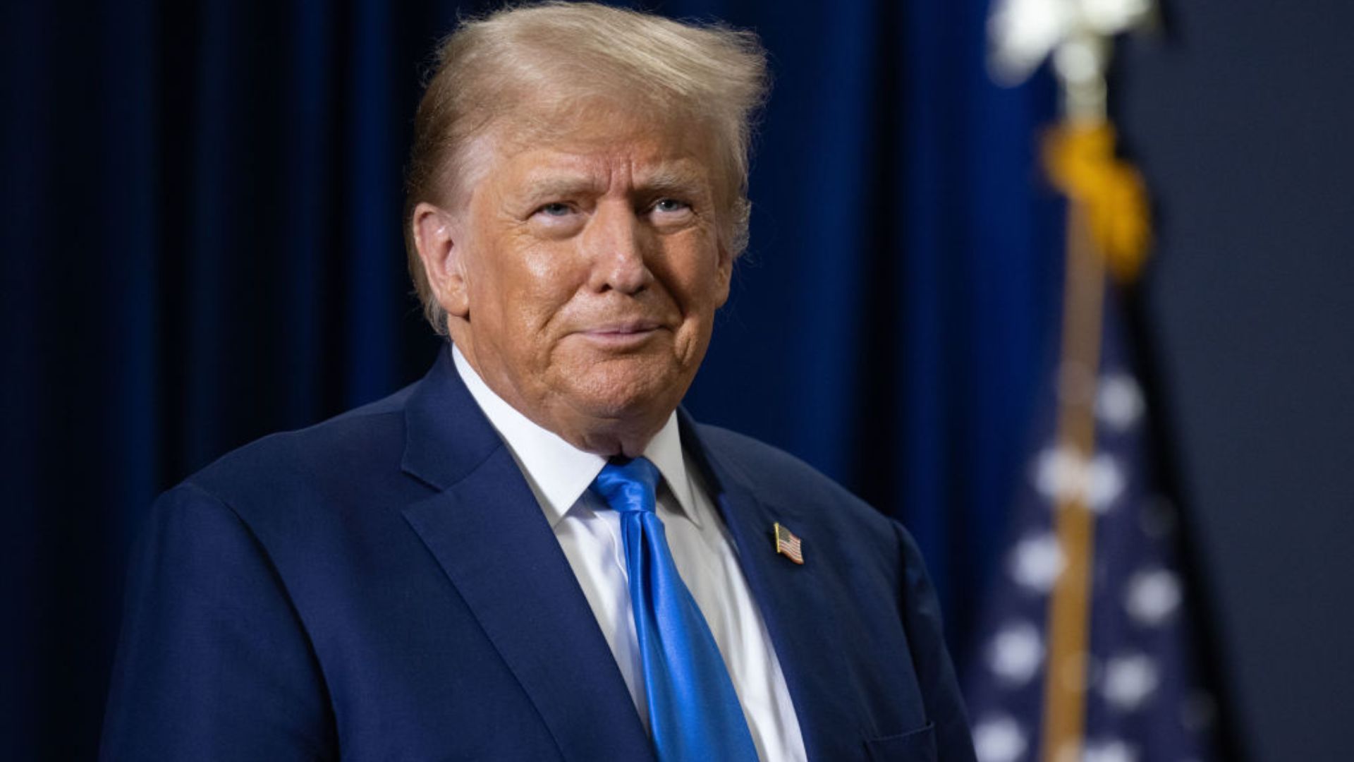 A close-up of Donald Trump wearing a dark suit and blue tie, looking slightly to his left. He stands in front of a dark blue curtain