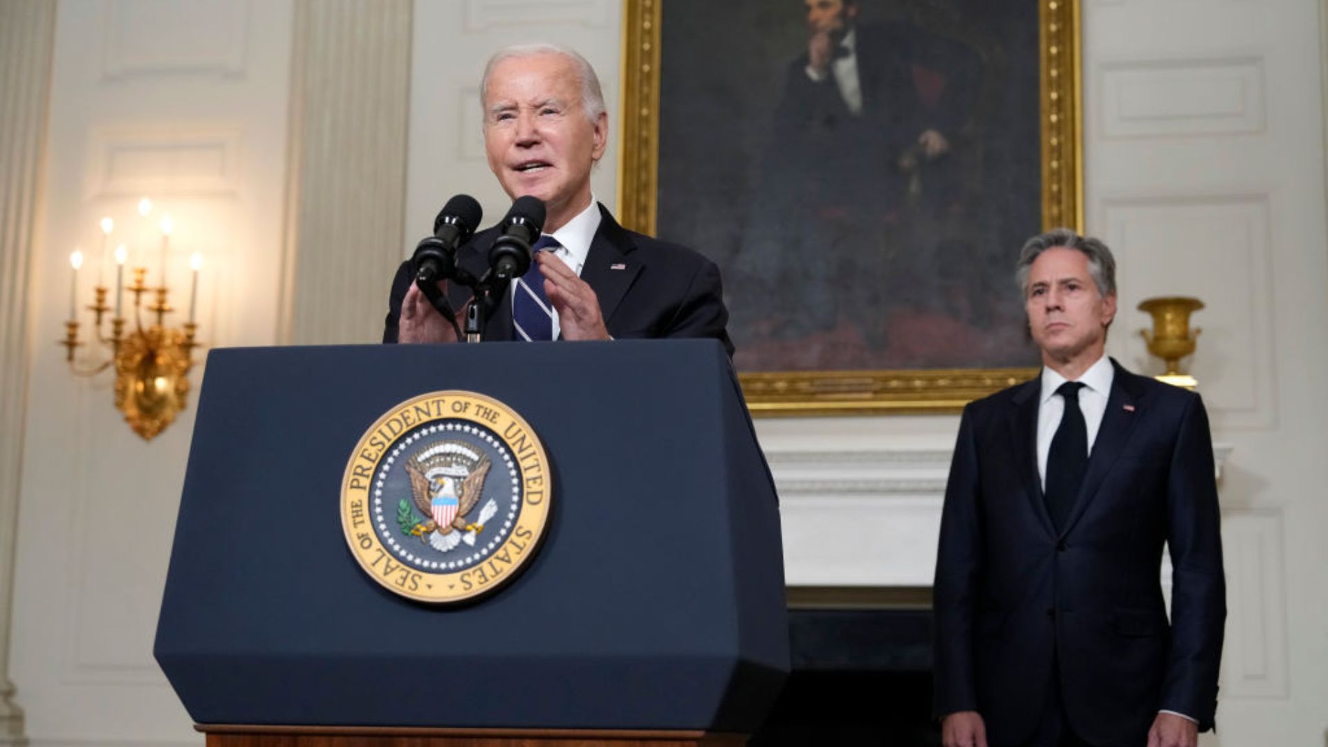 Biden stands behind a podium with a seal reading 'PRESIDENT OF THE UNITED STATES', speaking into microphones. Antony Blinken in a suit stands to the side, observing