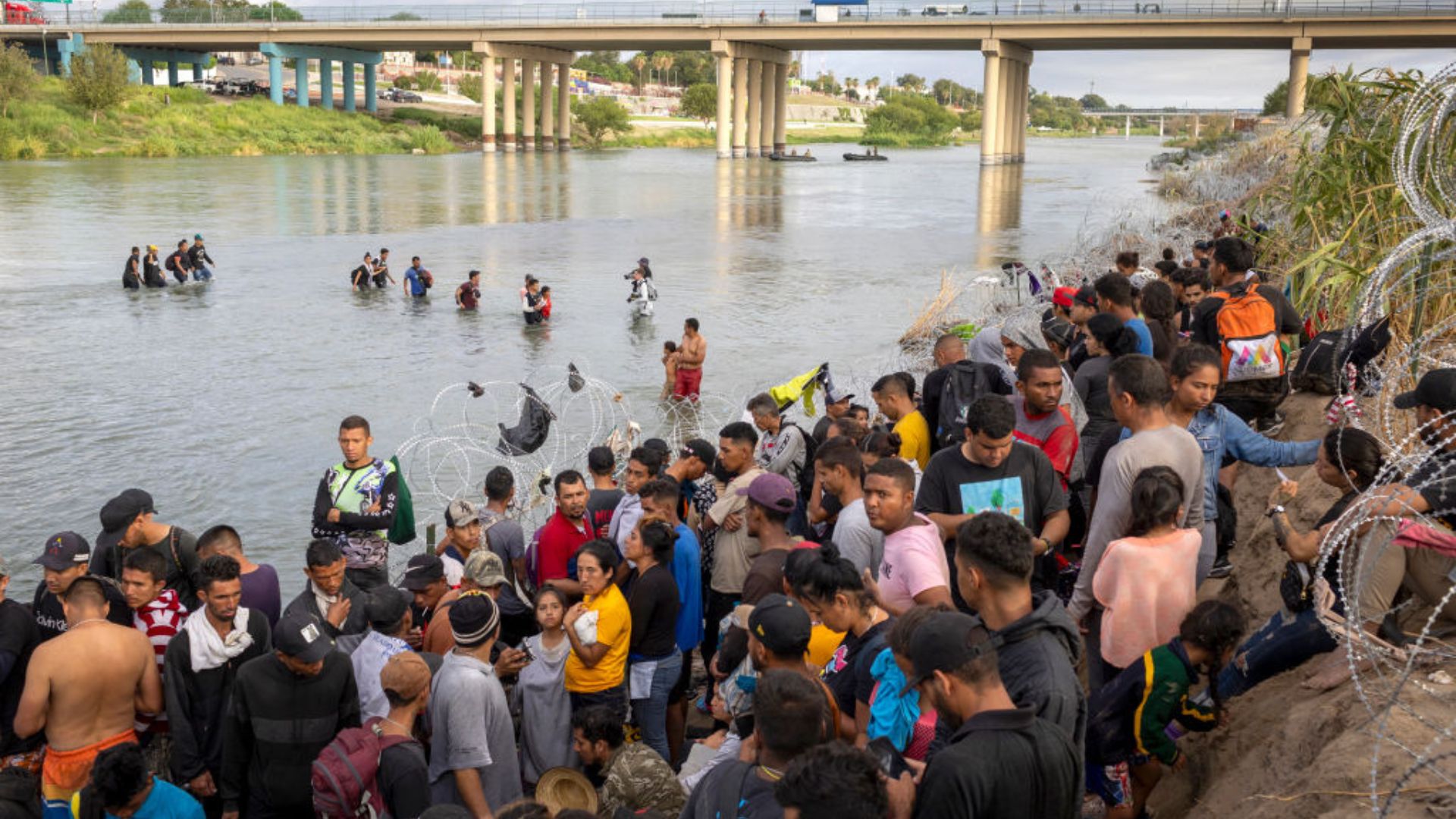A group of people gathered on the bank of a river with some wading through the water. In the background, a bridge spans the river. Many of the individuals are standing close to barbed wire barriers