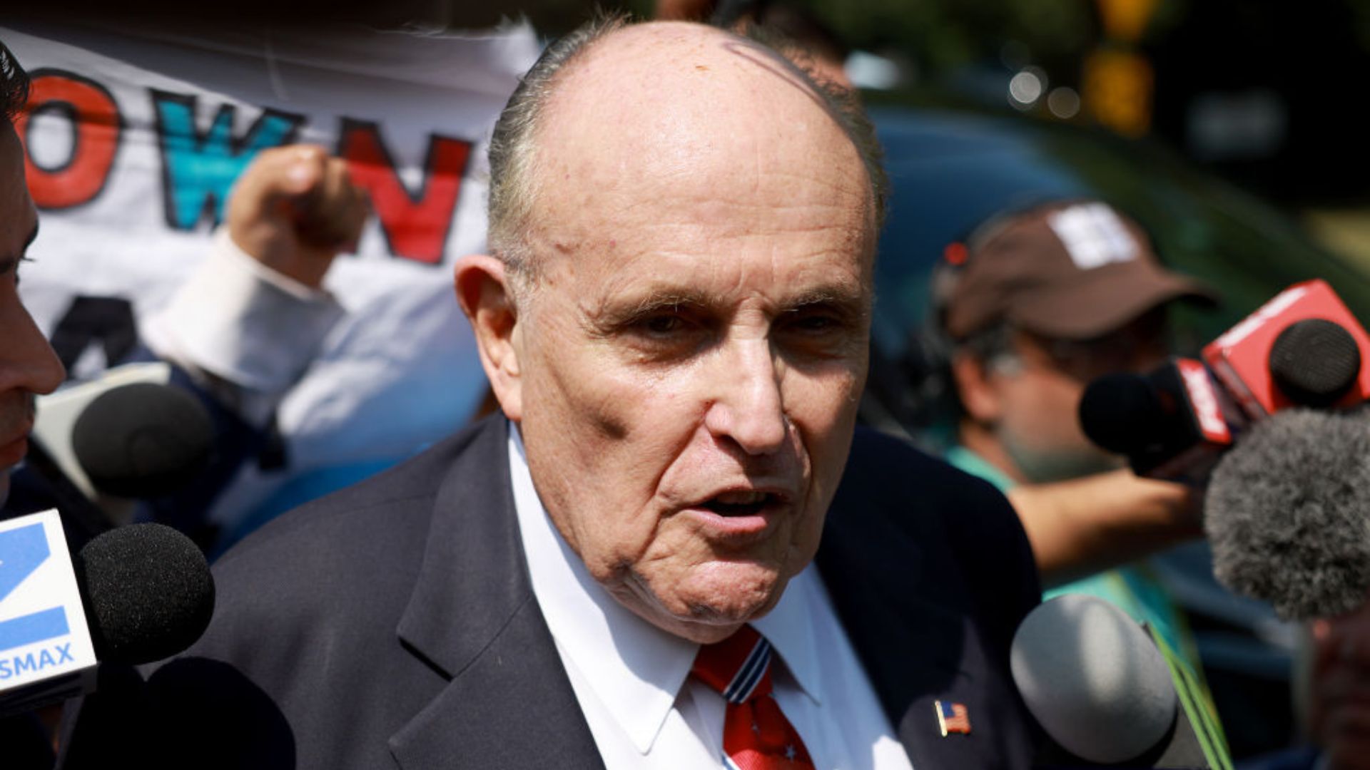 Rudy Giuliani, with a stern expression, addressing reporters outdoors. Microphones and cameras are pointed towards him. In the background, there's a blurred banner and various individuals
