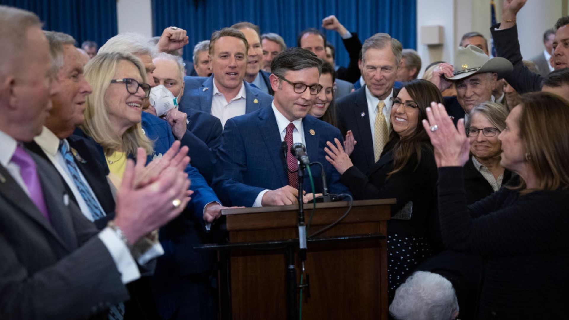 Rep. Mike Johnson, wearing glasses and a navy suit, stands at a podium speaking, surrounded by an enthusiastic group of individuals. Many are clapping, some raising their fists in support