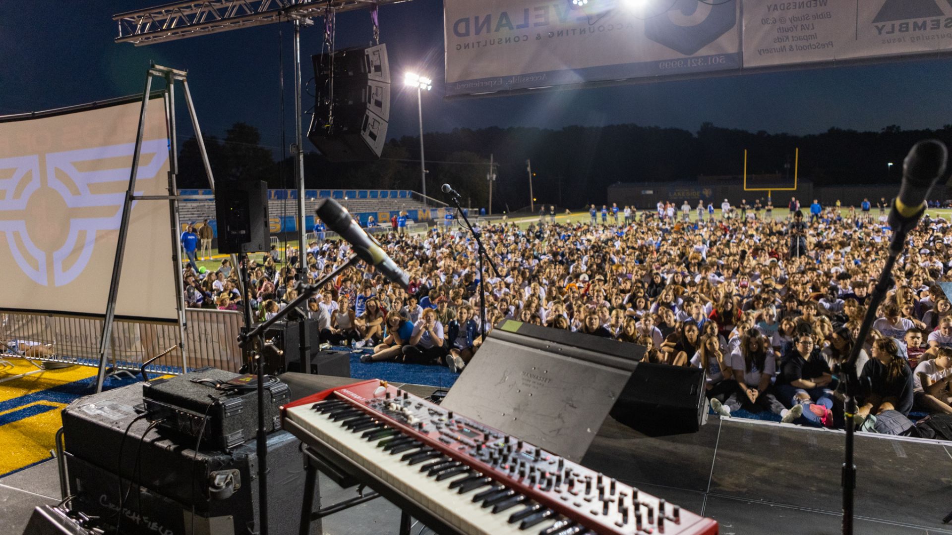 A view from the stage at an outdoor event, showing a large crowd of people seated on the field
