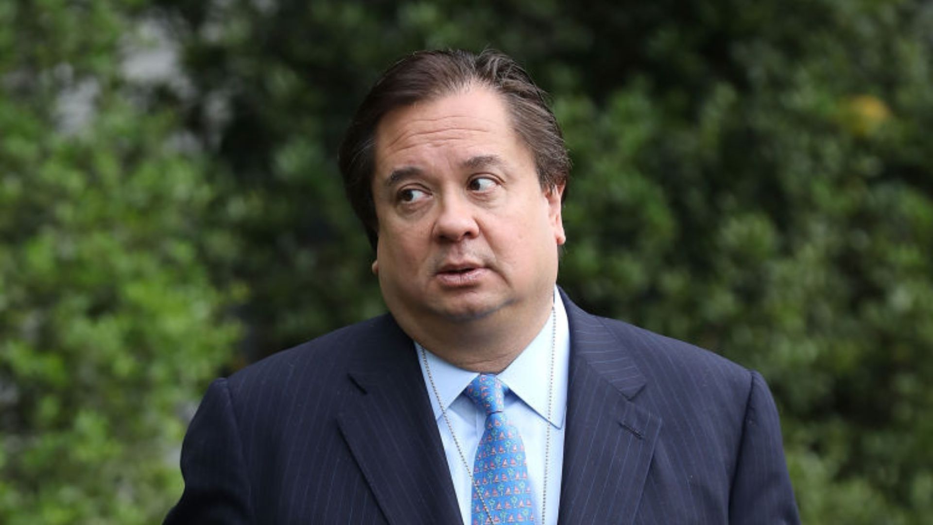 George Conway, wearing a dark suit and a patterned blue tie, stands outdoors with a backdrop of lush green trees