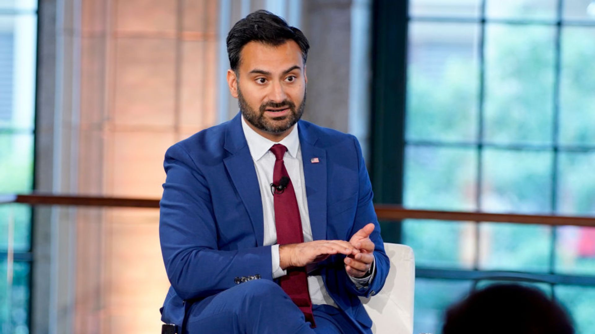 Ali Zaidi wearing a blue suit, red tie, and an American flag lapel pin, gestures while speaking