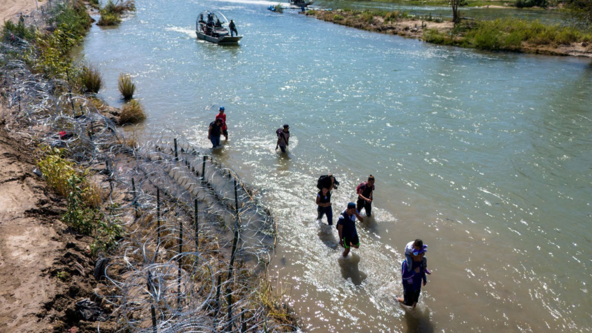Several people wading through a river with a backdrop of coiled barbed wire on the bank. A boat with two individuals is visible in the background