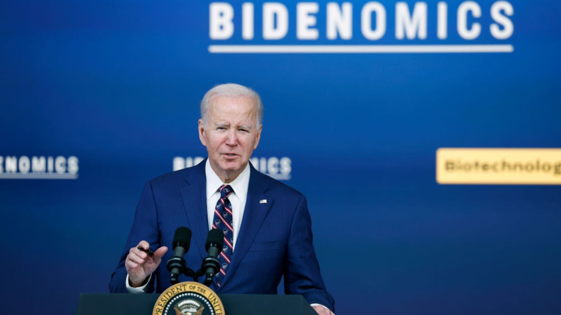 President Joe Biden standing on stage in a navy blue suit giving a speech in front of a ‘BIDENOMICS’ backdrop