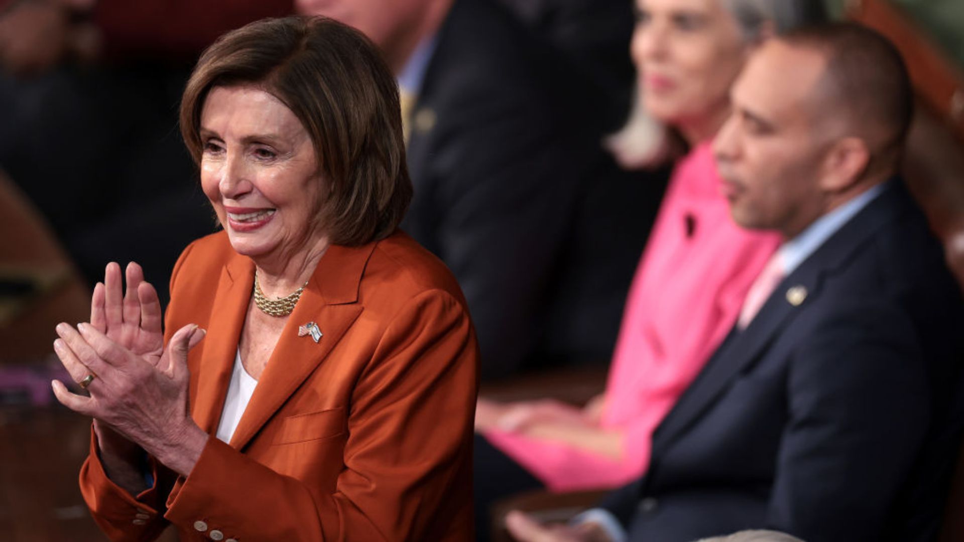 Nancy Pelosi wearing an orange blazer, smiling and clapping her hands, with blurred people in the background.