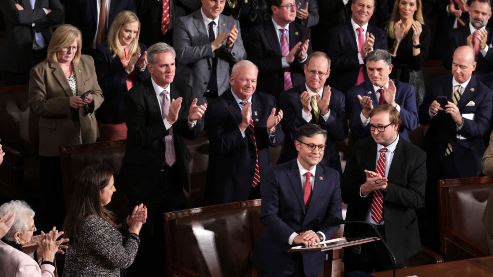 A diverse group of politicians standing and applauding Mike Johnson in a legislative chamber. Some are capturing the moment on their phones, while others are engaged in conversation