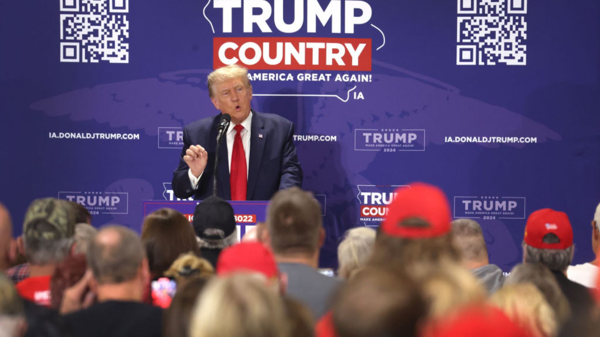 Trump addressing an audience from a podium, with a backdrop featuring logos, QR codes, and text banners