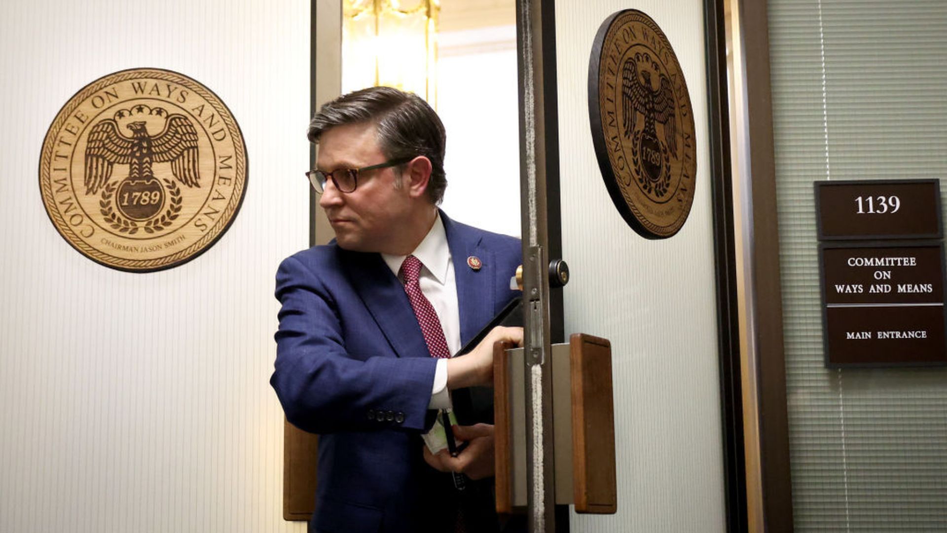 Mike Johnson, wearing a blue suit and red tie, opens a door with a wooden briefcase in hand
