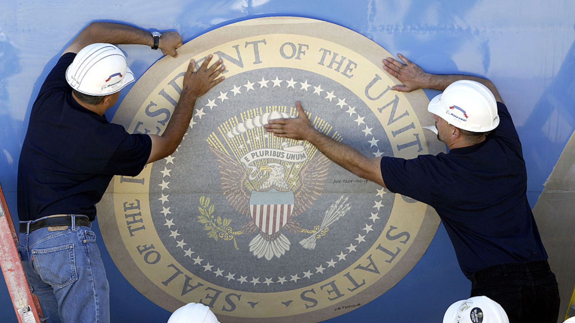 Two workers are carefully positioning and applying a large circular emblem displaying the Presidential Seal onto a blue surface