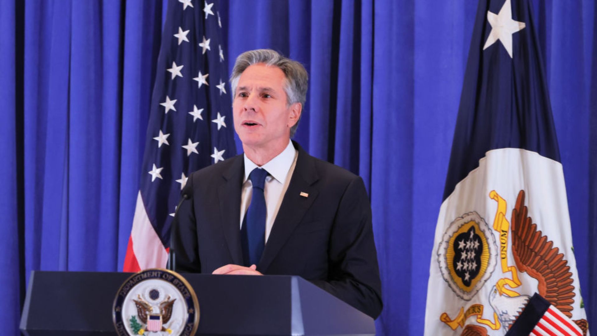 Antony Blinken, wearing a suit stands behind a podium with a seal reading 'DEPARTMENT OF STATE', speaking