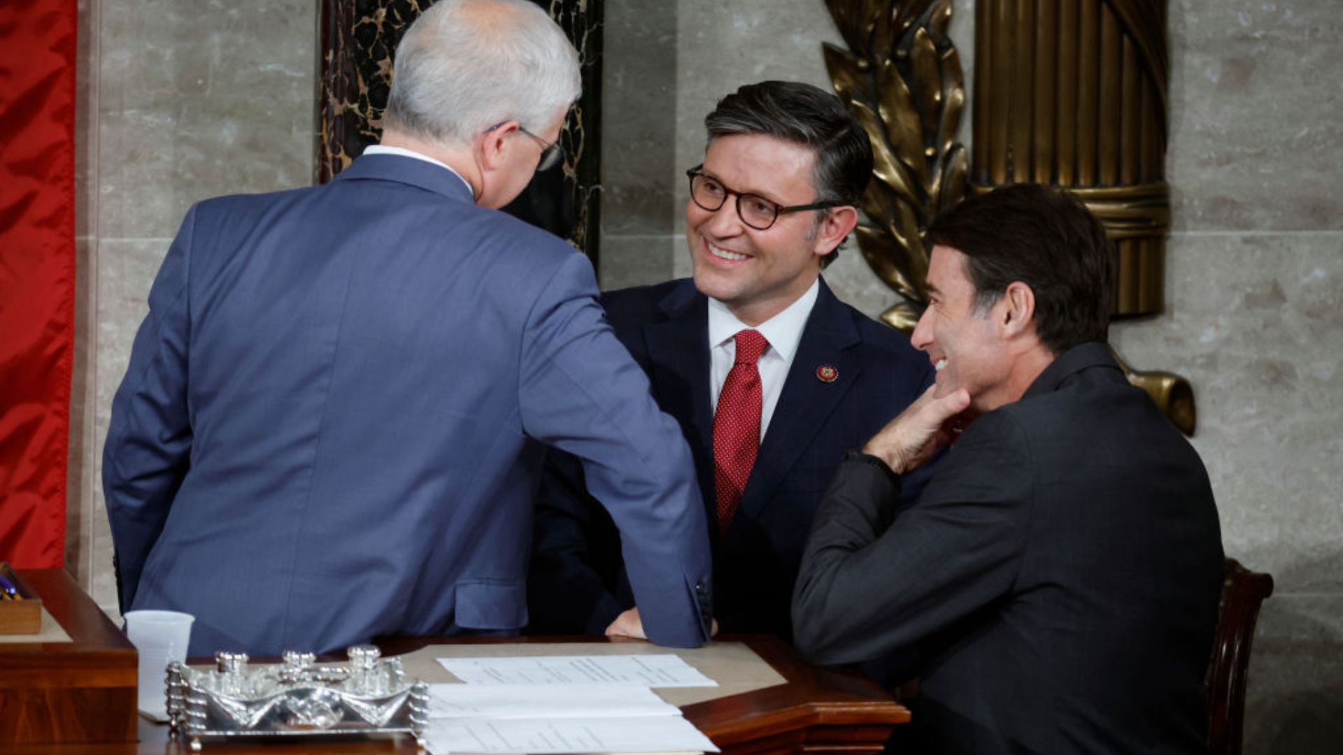Rep. Mike Johnson, wearing glasses and a navy suit with a red tie, smilingly interacts with an older man in a blue suit whose back is to the camera. Another individual in a black suit with a side profile is seen laughing to the right.