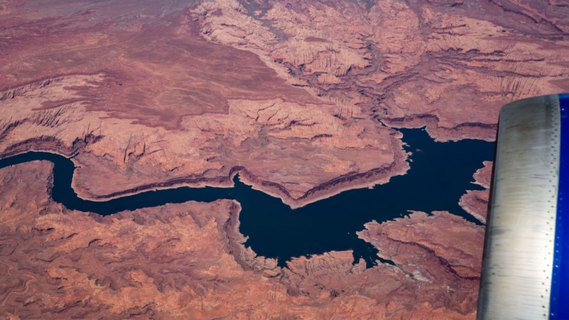 An aerial perspective of a deep blue water body winding through red and brown rugged terrain