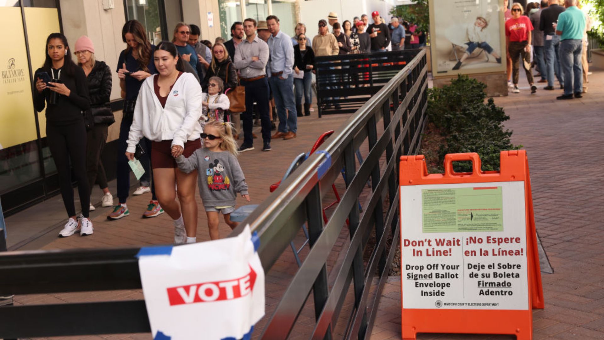A diverse group of individuals stands in line outside a building, waiting to vote, and in the foreground, an orange signboard offers the option to drop off signed ballots without waiting. The word 'VOTE' is prominently displayed on multiple signs