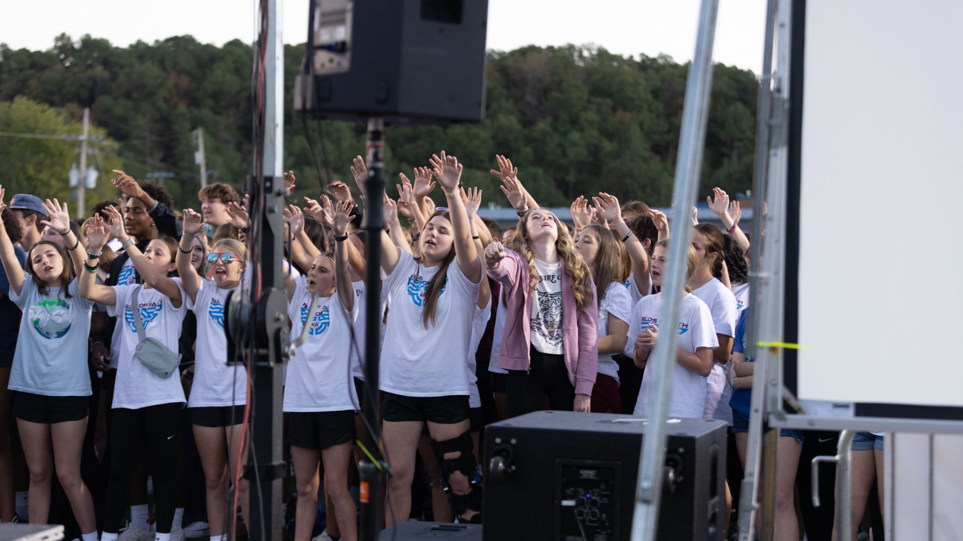 A diverse group of people, predominantly young individuals, enthusiastically raising their hands and engaging in an outdoor event. Many are dressed in white t-shirts with blue prints, and some are wearing sunglasses