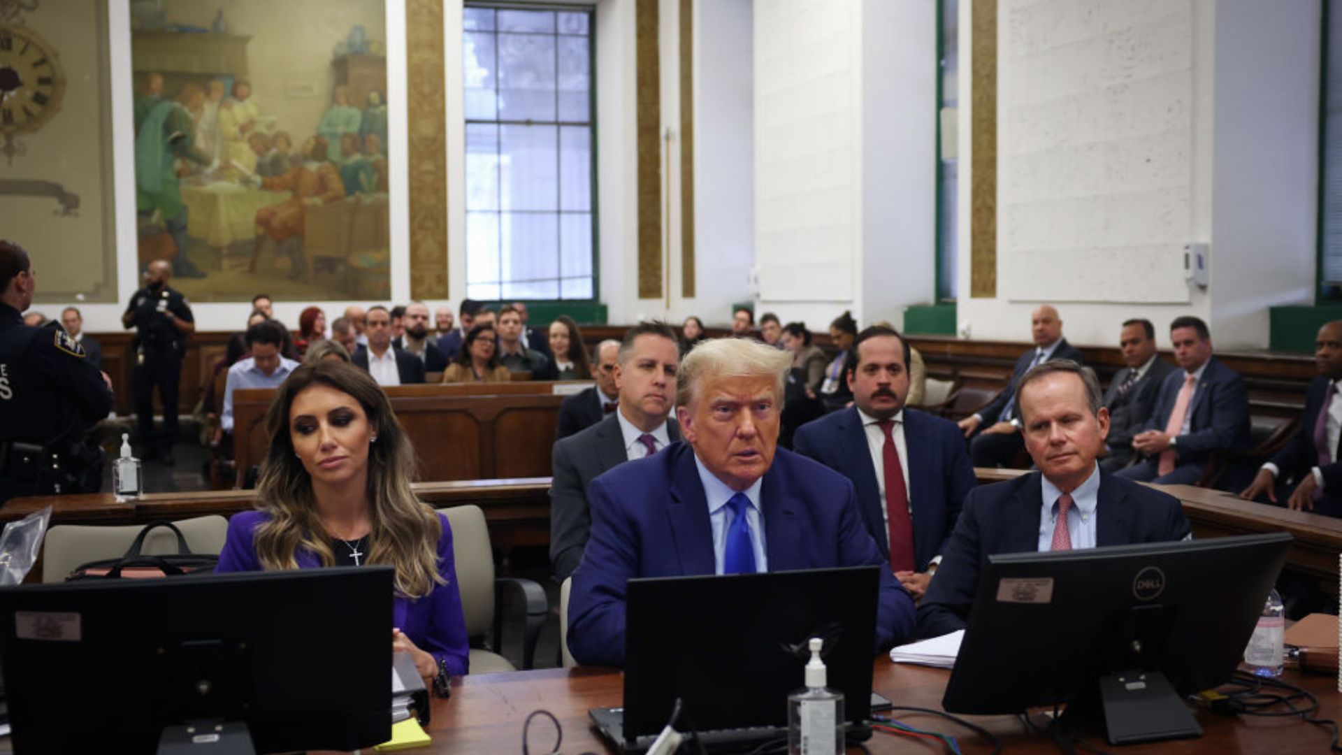 Former President Donald Trump seated in a courtroom alongside other individuals, appearing focused on the proceedings