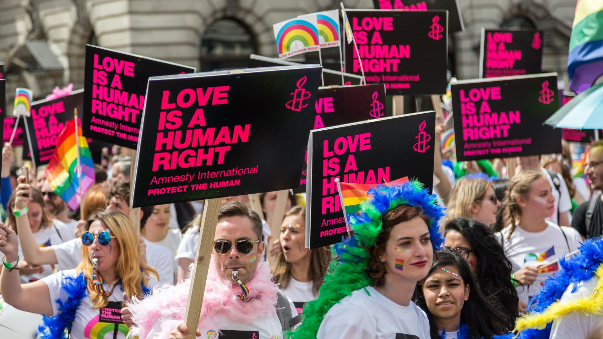 A group of people at a pride parade, holding up black signs that read "LOVE IS A HUMAN RIGHT - Amnesty International - PROTECT THE HUMAN"