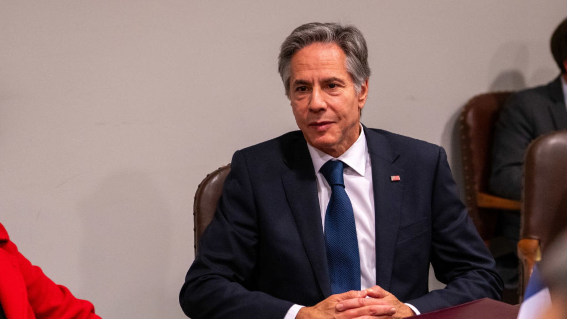 Antony Blinken in a navy blue suit with a light blue tie sits with his hands clasped, attentively participating in a discussion