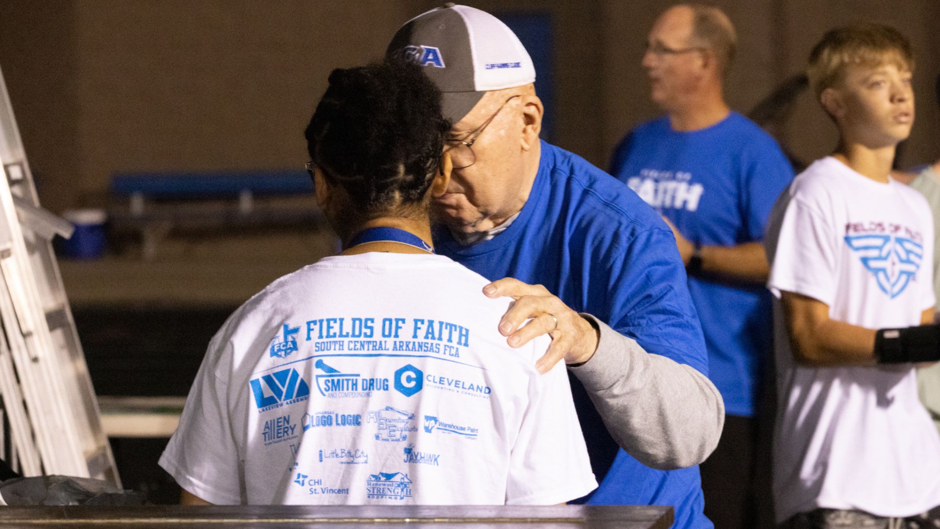 Two individuals at an outdoor event, where an older man in a blue shirt and cap is embracing a person wearing a white t-shirt with multiple logos and the text "FIELDS OF FAITH"