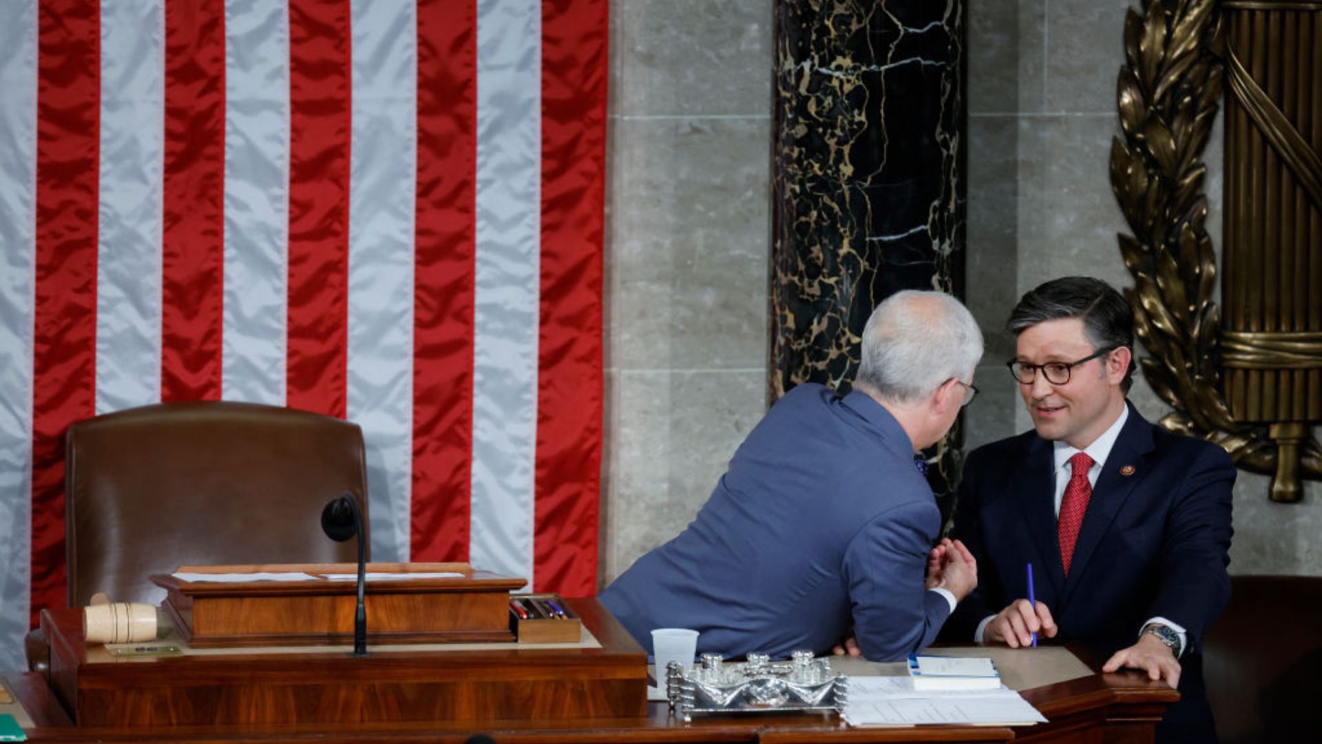 Mike Johnson, wearing glasses and a dark suit with a red tie, converses with another individual in a blue shirt in front of a large American flag backdrop