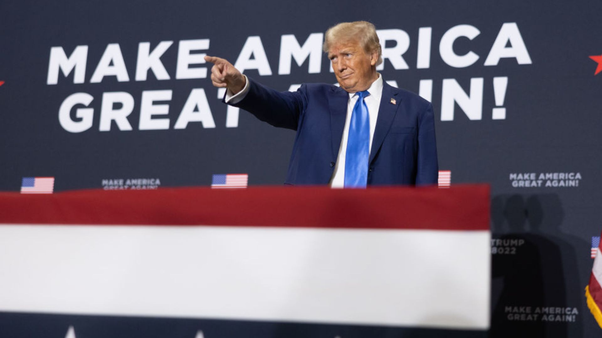 Donald Trump, wearing a dark suit and blue tie, points towards his right while standing in front of a backdrop with the slogan "MAKE AMERICA GREAT AGAIN!"