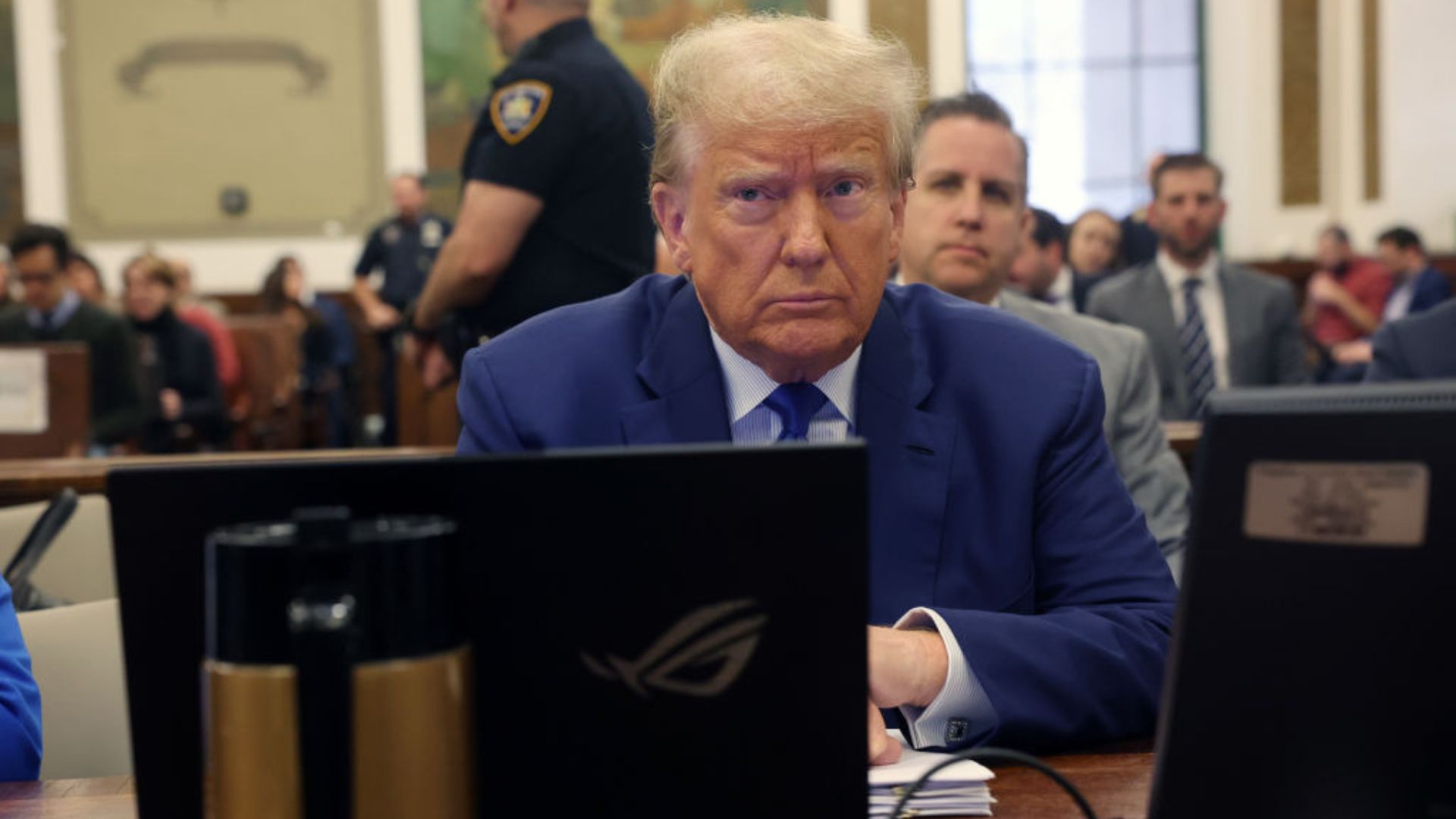 Donald Trump, wearing a blue suit, sits at a table in a room with a laptop in front of him