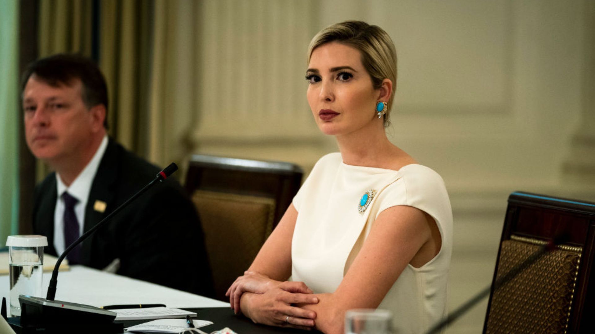 Ivanka Trump, wearing a white dress adorned with a turquoise brooch, sits at a table