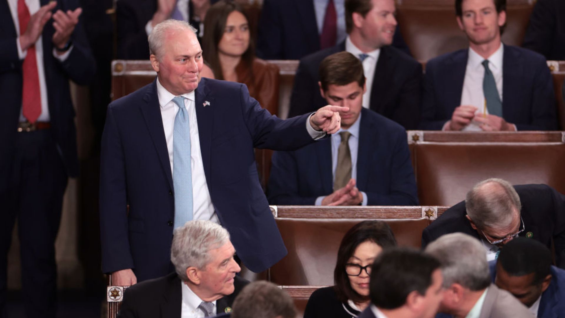 Steve Scalise in a blue suit and light blue tie, pointing to something in the distance, surrounded by other individuals in a formal setting with wooden benches