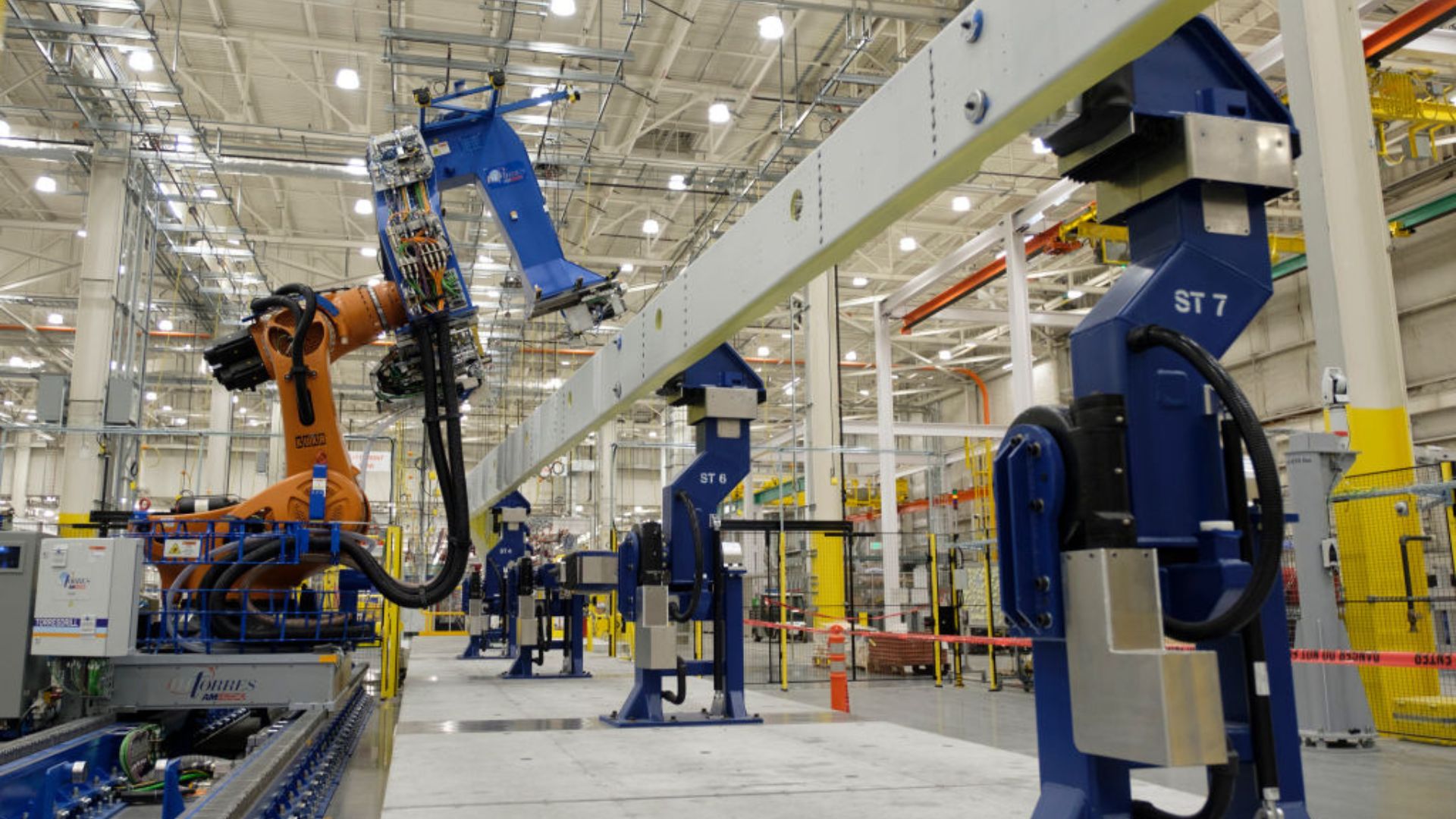 A spacious industrial warehouse with high ceilings, illuminated by overhead lights. In the foreground, an orange robotic arm with wiring and tubing stands next to blue machinery