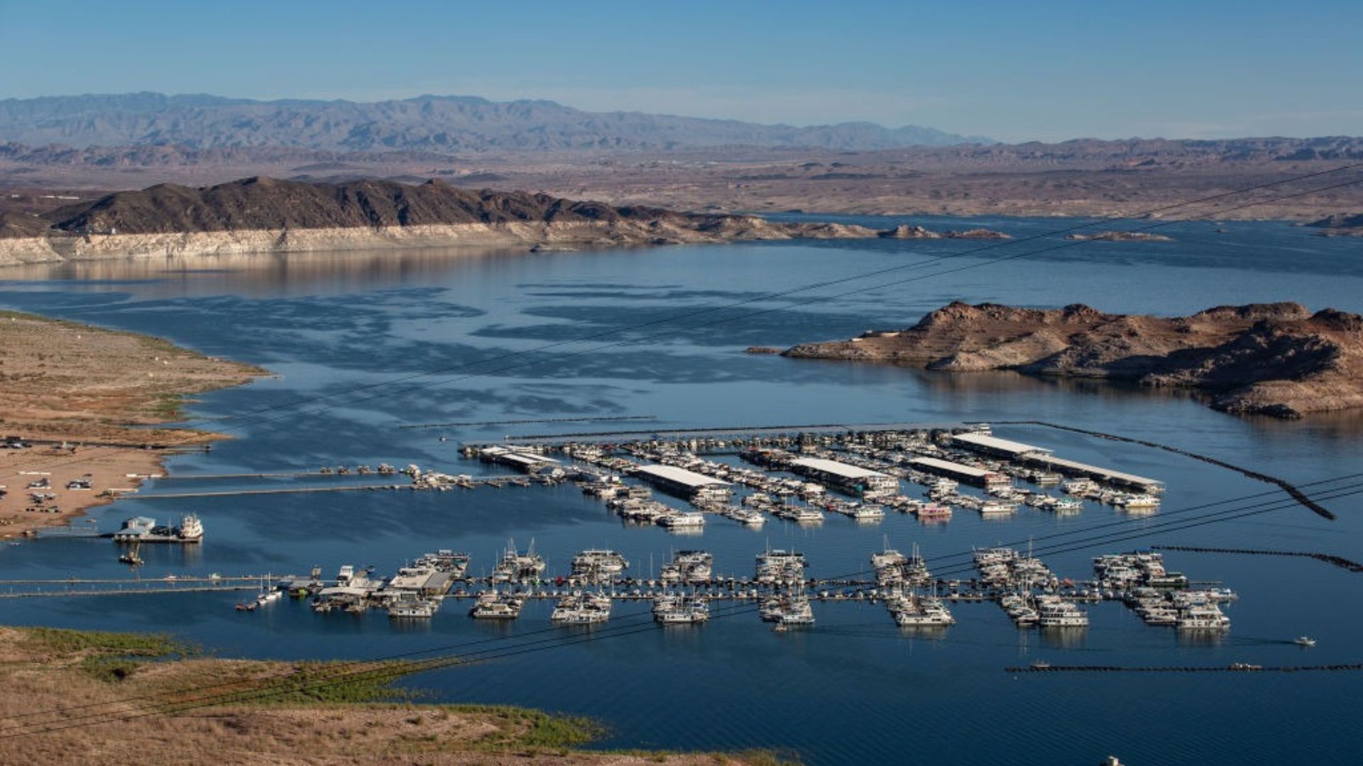 An aerial view of a large marina filled with moored boats. The marina is situated on a vast lake surrounded by rugged terrain and mountains in the distance