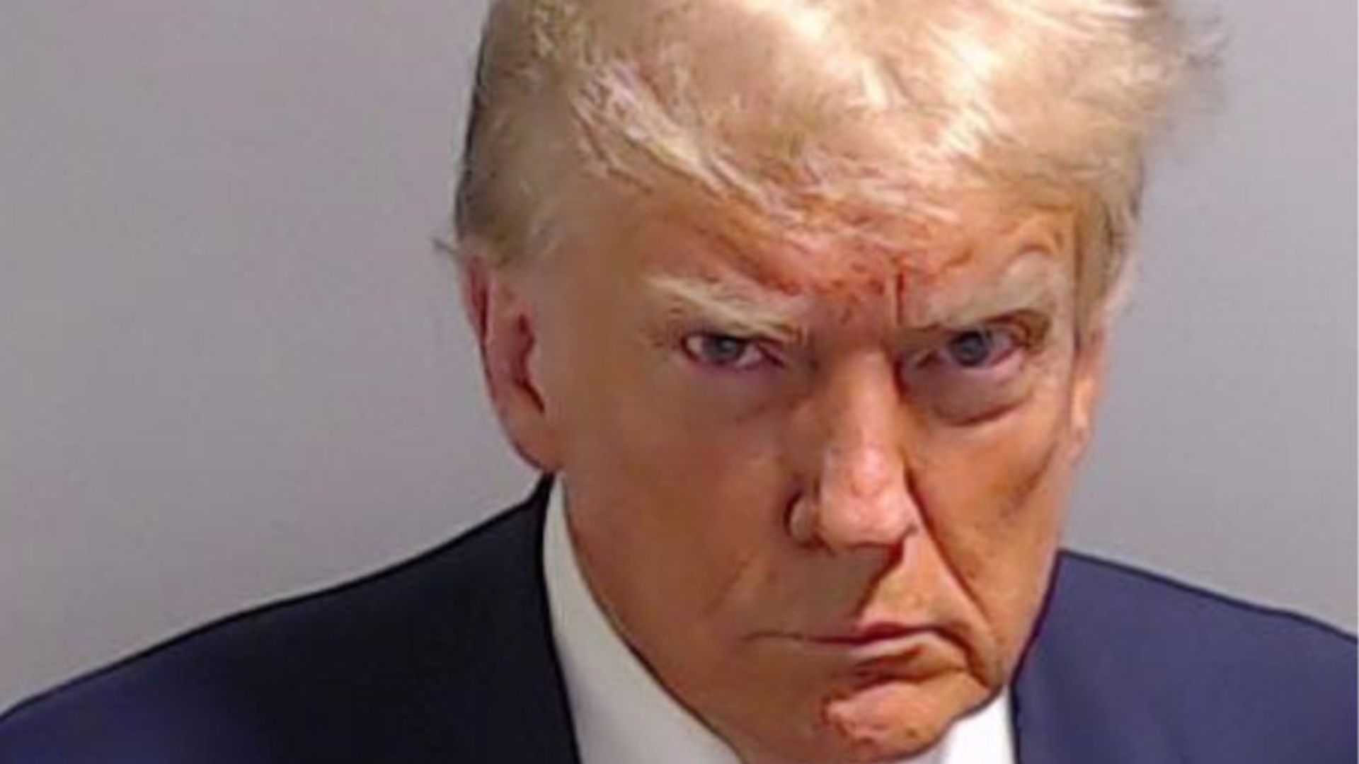 Close-up of Donald Trump's face, looking directly at the camera with a stern expression. The image has a neutral background, and he is wearing a dark suit